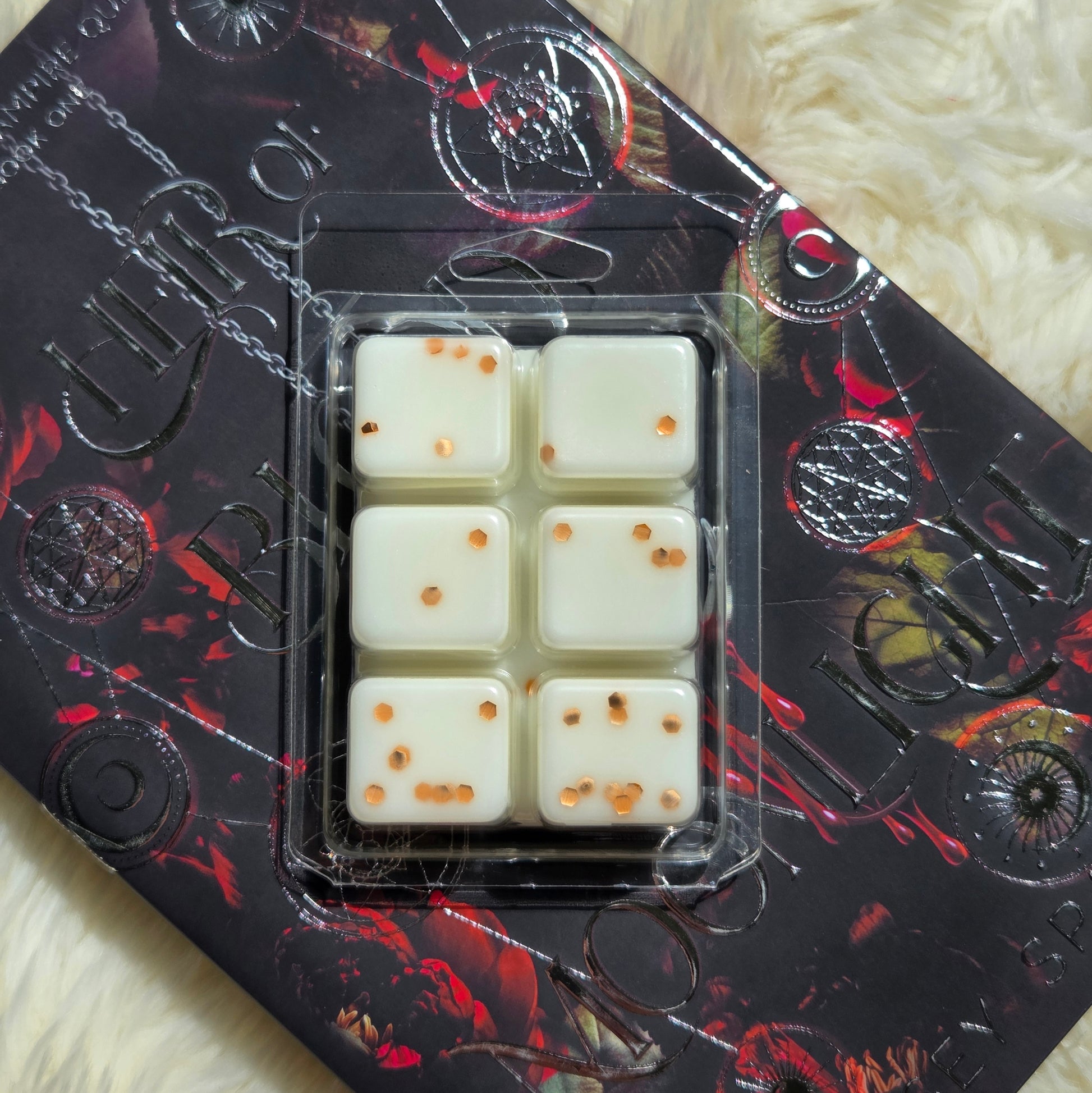 Six square cream-colored wax melts cubes with copper eco-friendly glitter, packaged in a clear plastic clamshell, resting on a black and red gothic-themed decorative book over a white faux fur background.