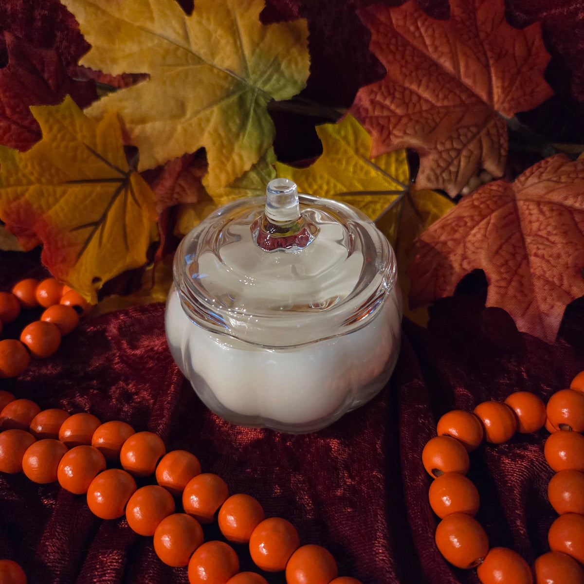 A small clear glass pumpkin-shaped jar with a matching lid and white wax inside, styled on dark red velvet fabric with orange beads and autumn leaves around it.