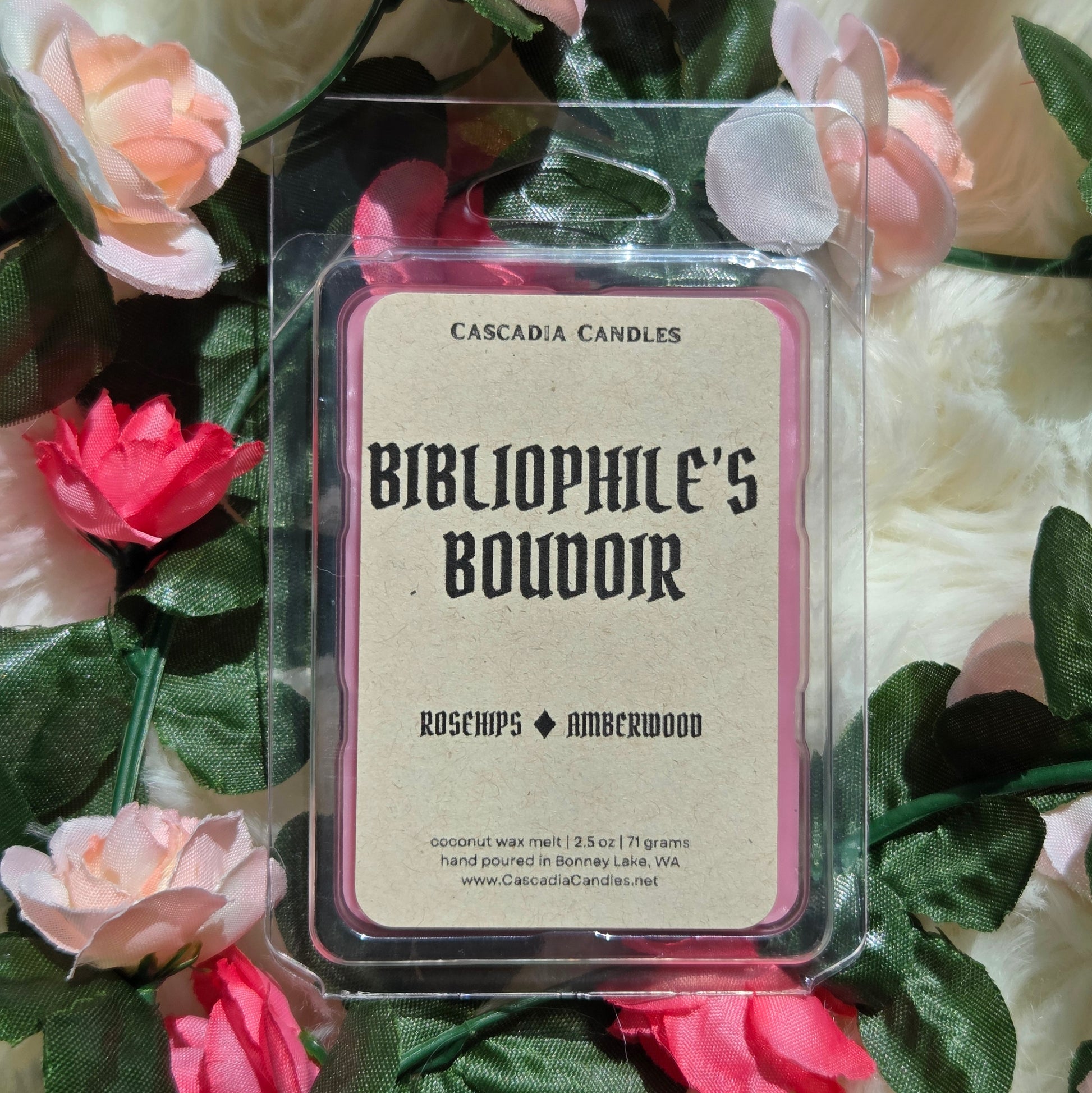 A packaged wax melt from Cascadia Candles labeled 'Bibliophile's Boudoir' in a clear plastic clamshell, surrounded by pink and peach roses and green leaves on a soft cream faux fur background.