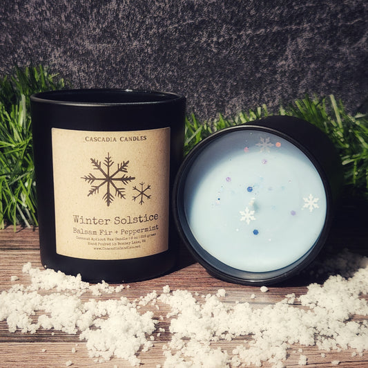 Cascadia Candles Winter Solstice candle with a black lid on a wooden surface with snowflakes.