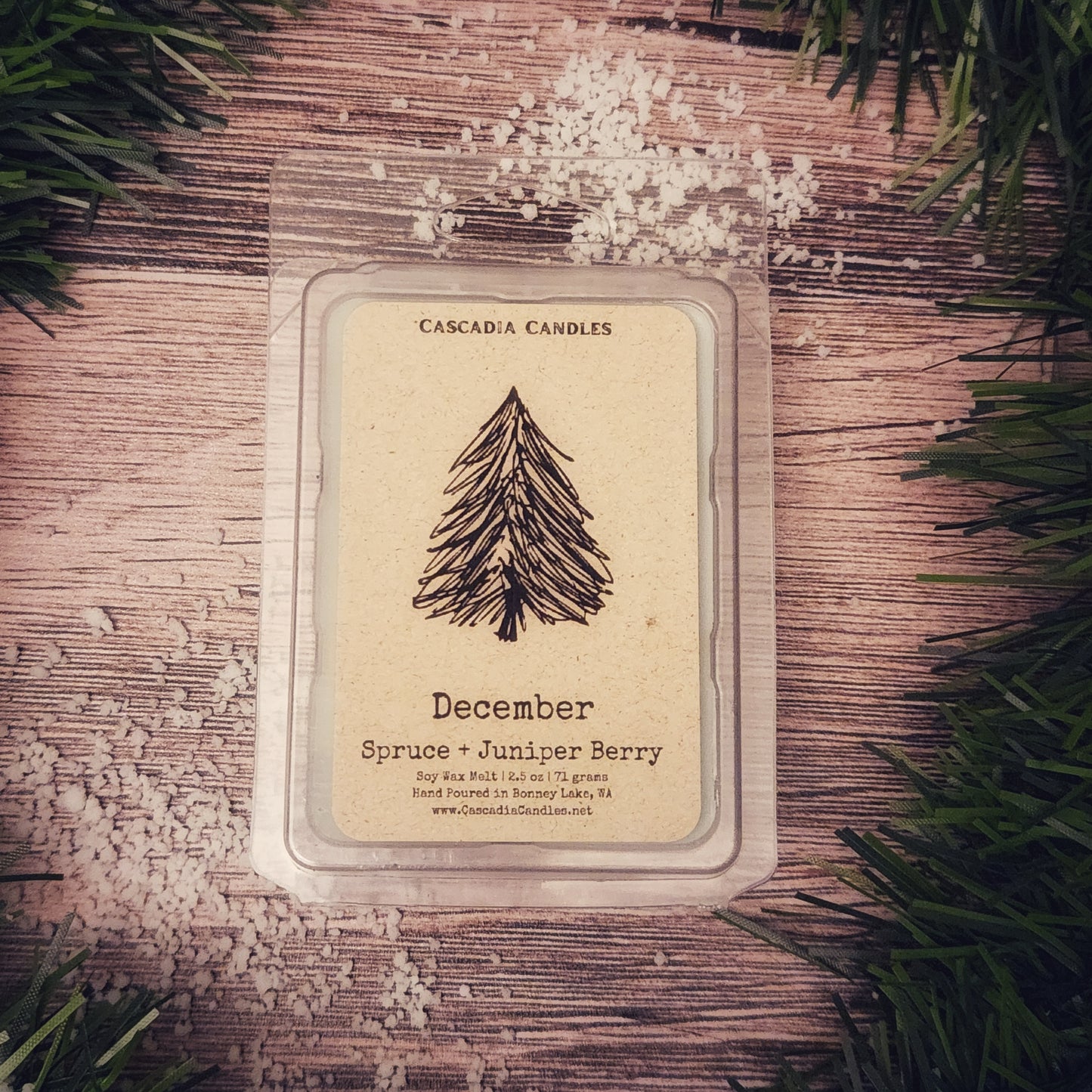 Cascadia Candles December Spruce + Juniper Berry candle on a wooden surface with greenery around.