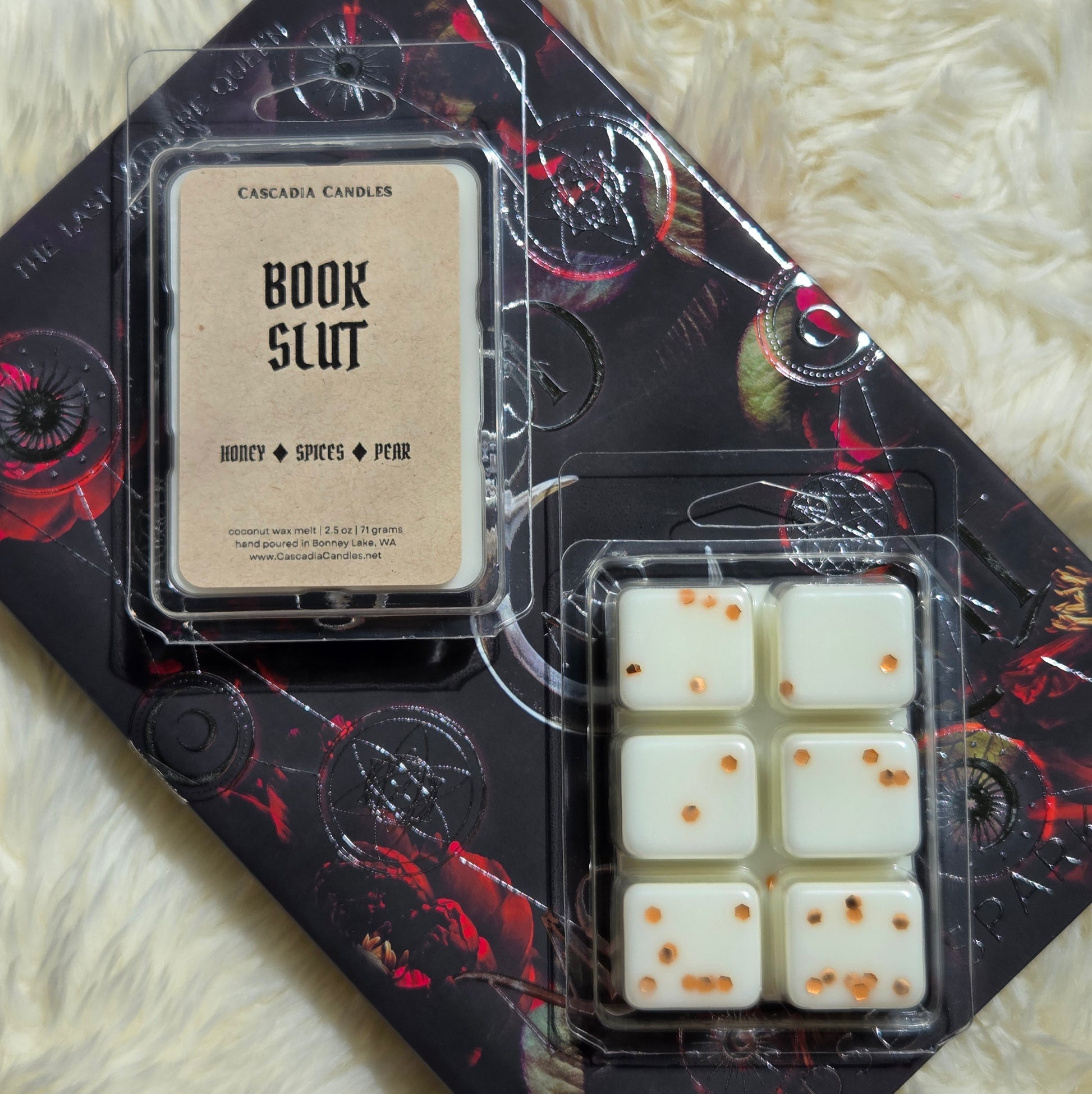 Cascadia Candles Book Slut wax melt set, featuring a Kraft paper label and a clear plastic clamshell of six white wax melts with copper eco-friendly glitter, displayed on a black and red patterned book over a white faux fur background.