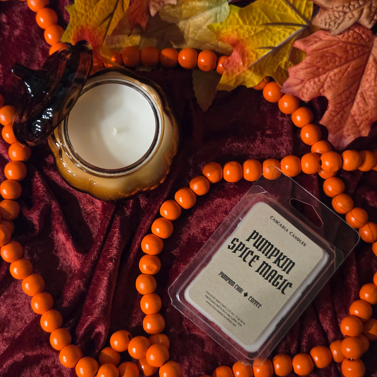 A wax melt package labeled 'Pumpkin Spice Magic' by Cascadia Candles, styled in a flat lay with orange beaded garland, autumn leaves, and a pumpkin-shaped candle on a burgundy velvet background.