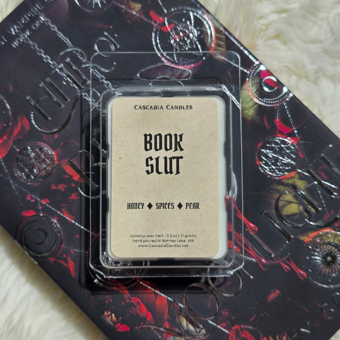 A packaged Cascadia Candles wax melt labeled 'Book Slut' resting on top of a book. The wax melt package is clear plastic with a Kraft paper label, and the book underneath has a dark design with red and black artwork and embossed circular motifs.