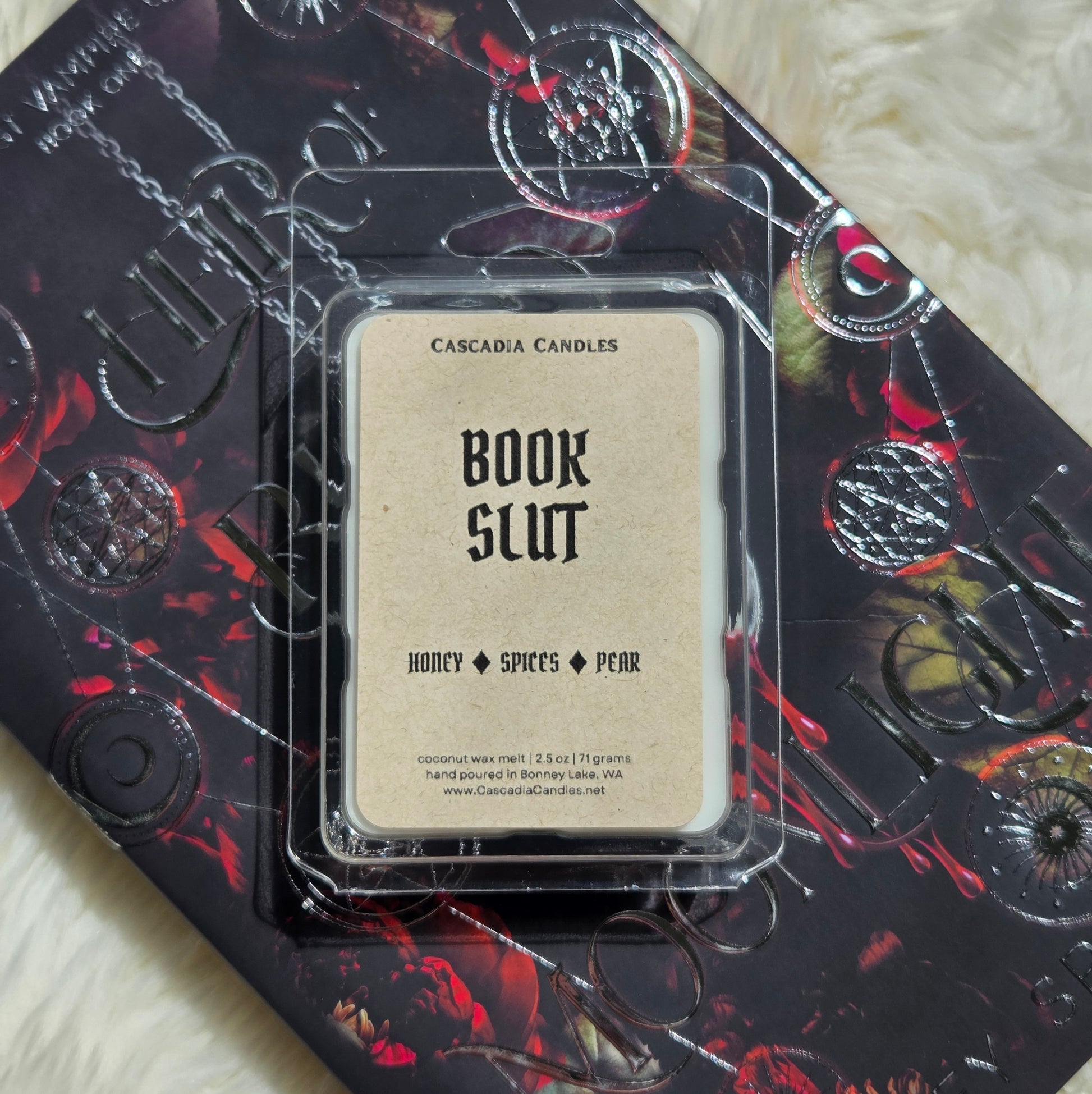 A packaged Cascadia Candles wax melt labeled 'Book Slut' resting on top of a book. The wax melt package is clear plastic with a Kraft paper label, and the book underneath has a dark design with red and black artwork and embossed circular motifs.