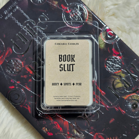 A packaged Cascadia Candles wax melt labeled 'Book Slut' resting on top of a book. The wax melt package is clear plastic with a Kraft paper label, and the book underneath has a dark design with red and black artwork and embossed circular motifs.