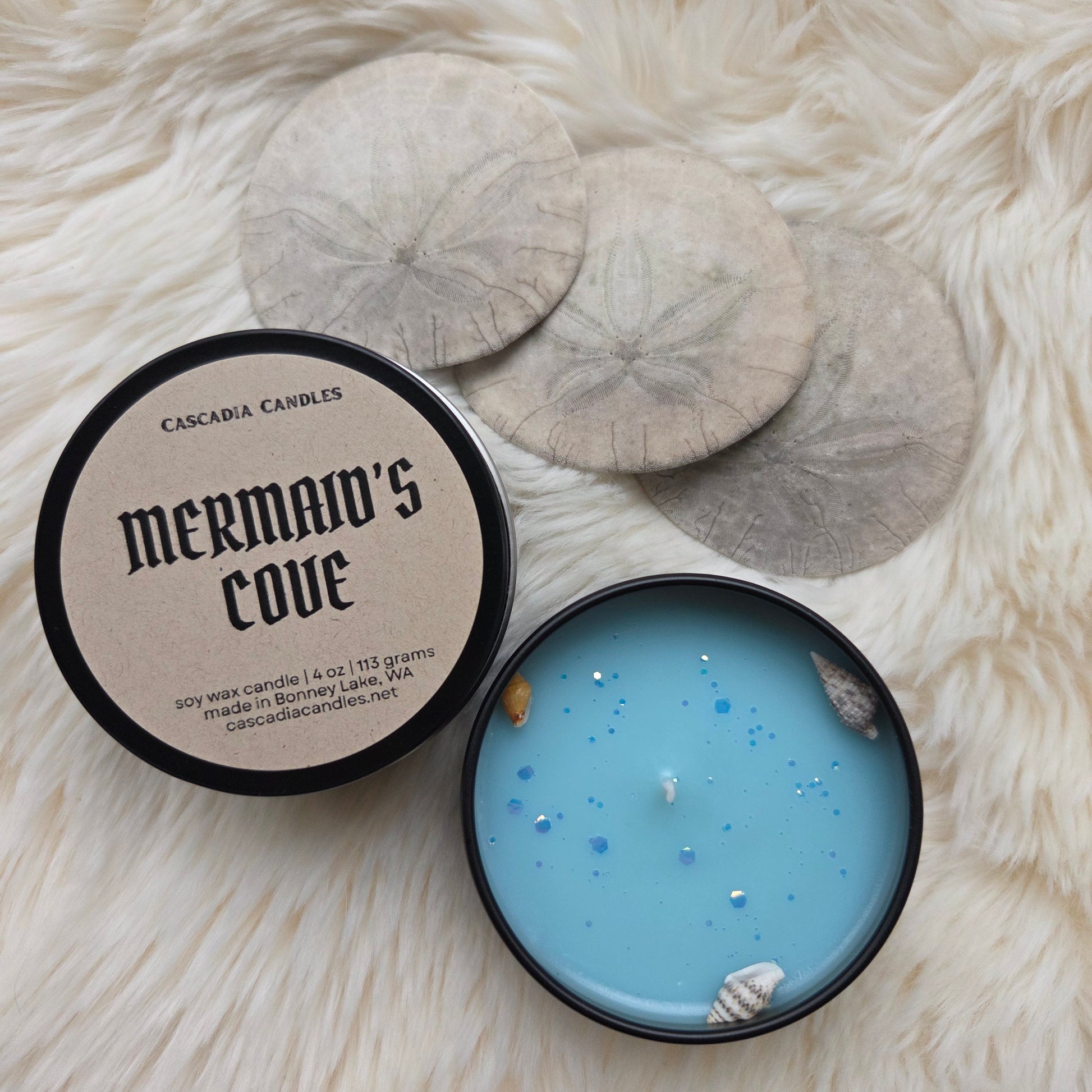 Blue candle in a black container with 'Mermaid's Cove' branding on a fluffy white surface.