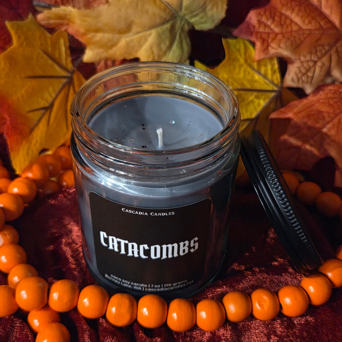 An open jar candle labeled Cascadia Candles 'Catcombs' sits on a dark red fabric surrounded by orange beads and autumn leaves. The candle wax is a muted gray-blue, and the lid rests beside the jar.