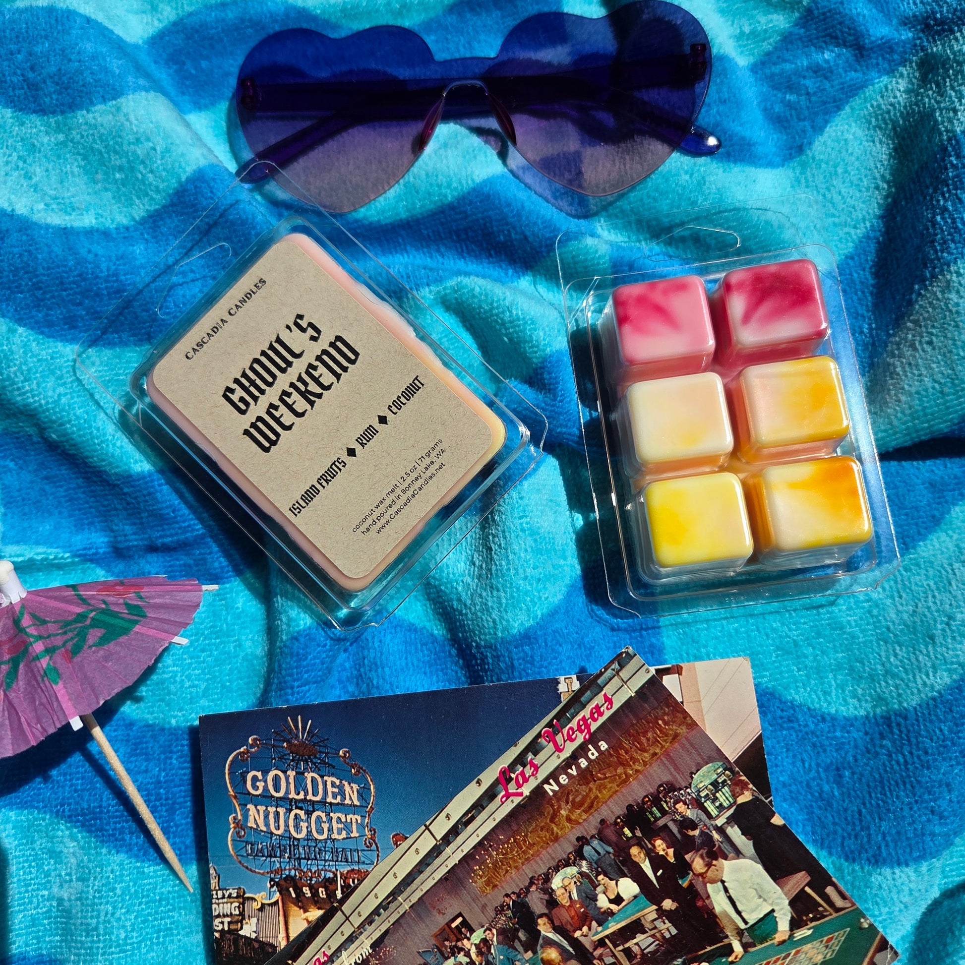 Two wax melts packages on a blue textured surface with sunglasses and postcards.