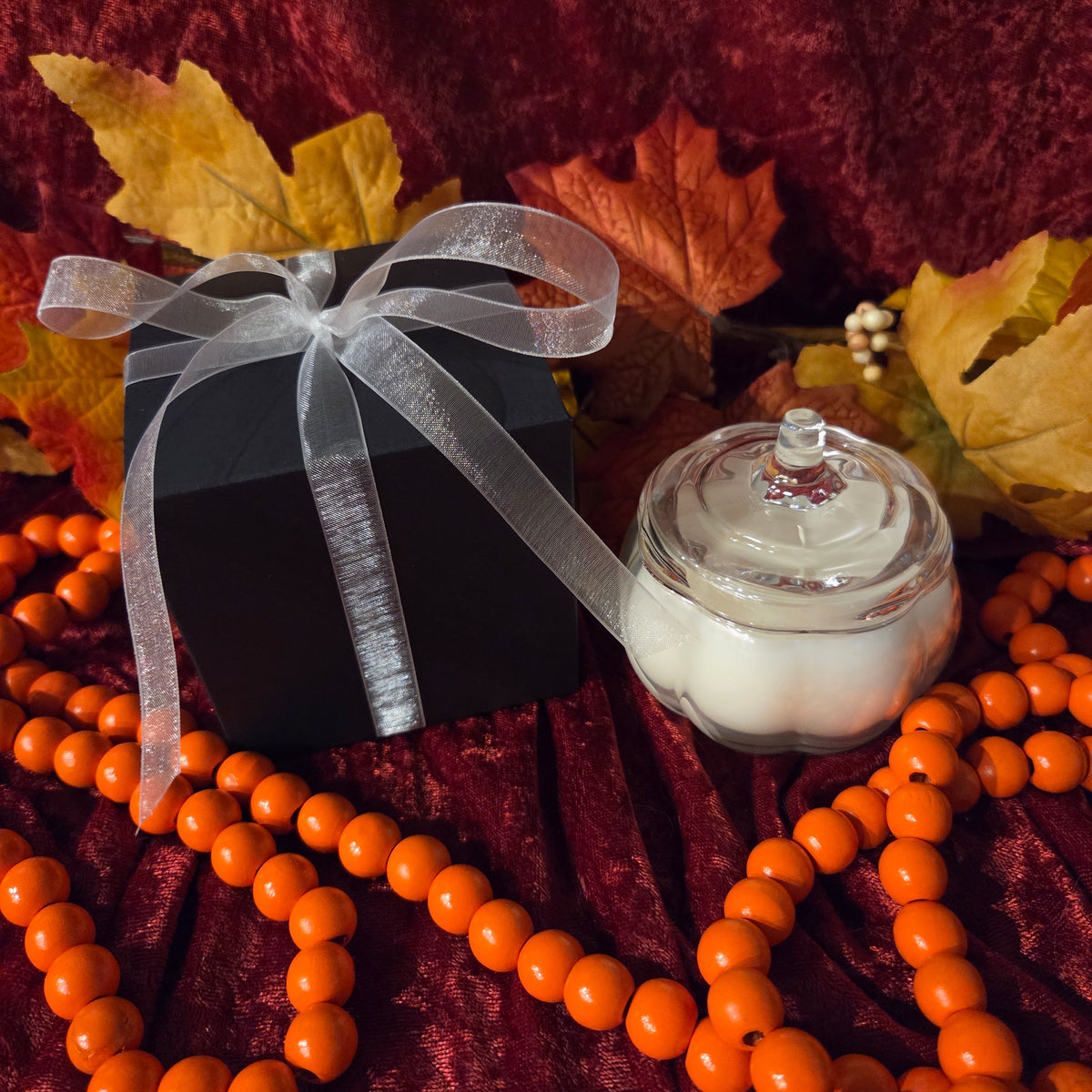 A black gift box tied with a sheer white ribbon sits beside a small clear glass pumpkin-shaped jar filled with white wax, styled with orange bead garlands, autumn leaves, and a dark red fabric backdrop.