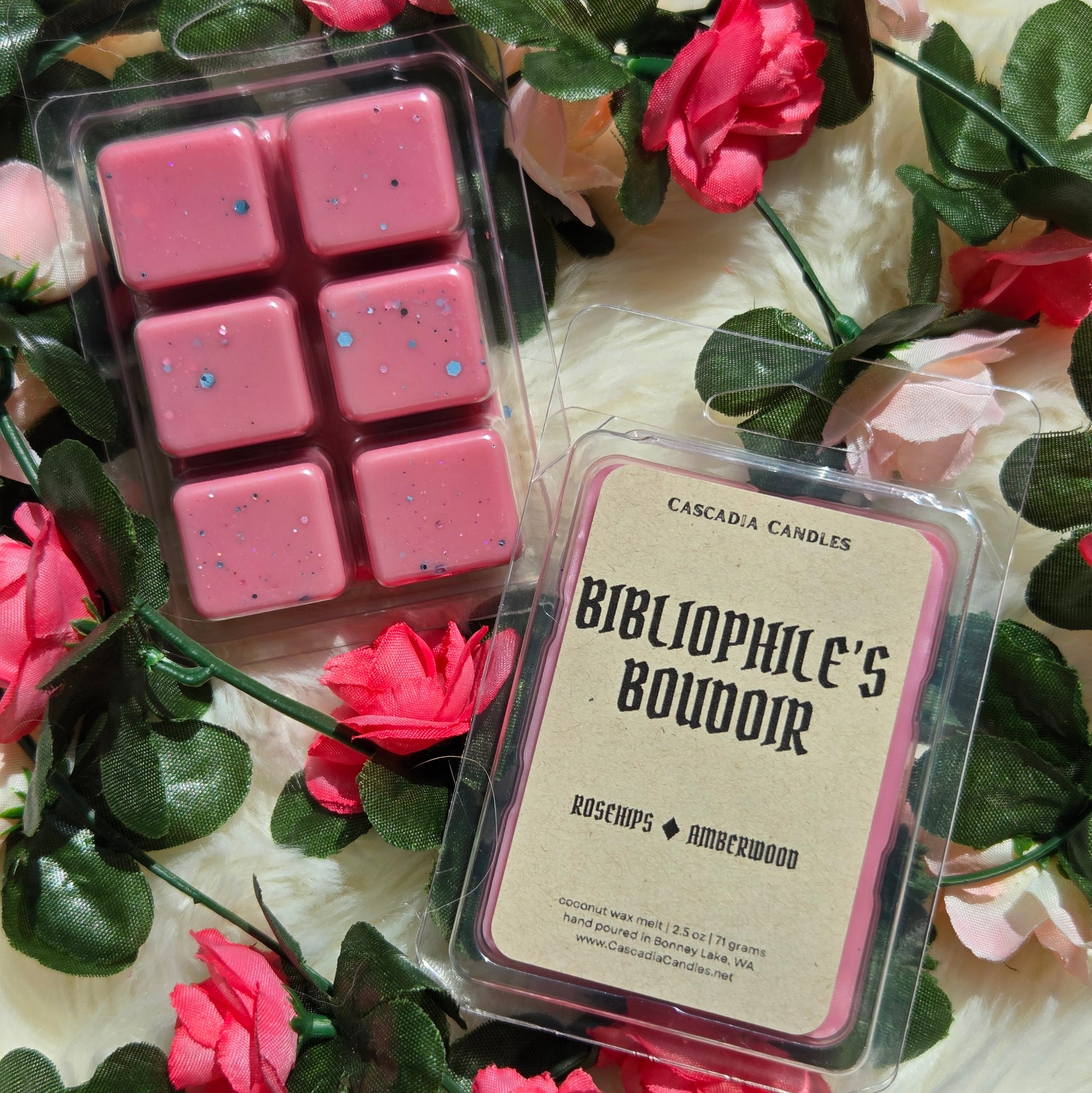 Two packaged wax melts from Cascadia Candles are arranged on a white faux-fur surface with pink roses and green leaves. One package contains six pink wax cubes with eco-friendly glitter, and the other shows a front label reading 'BIBLIOPHILE'S BOUDOIR' with rose and amberwood notes.