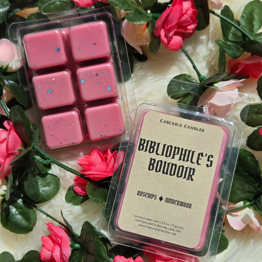 Two packaged wax melts from Cascadia Candles are arranged on a white faux-fur surface with pink roses and green leaves. One package contains six pink wax cubes with eco-friendly glitter, and the other shows a front label reading 'BIBLIOPHILE'S BOUDOIR' with rose and amberwood notes.