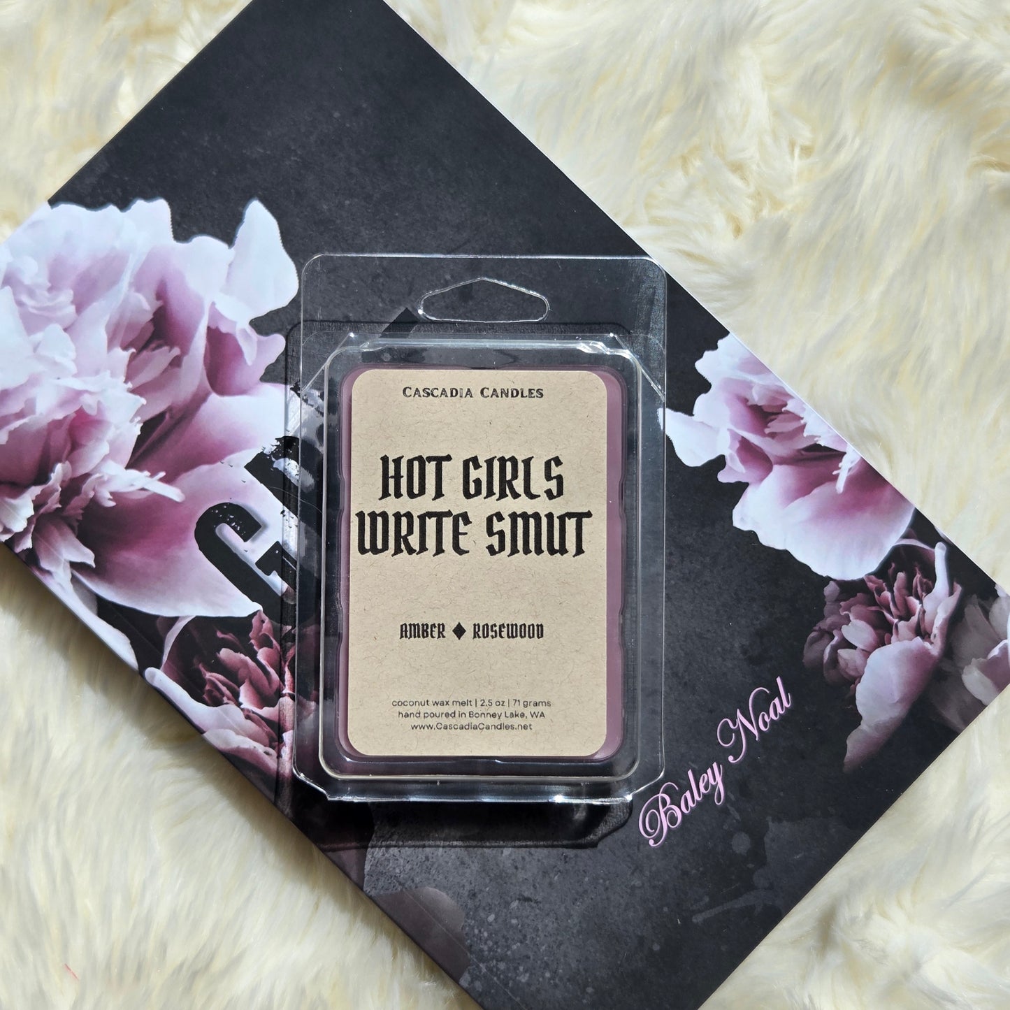 A packaged wax melt product labeled 'HOT GIRLS WRITE SMUT' by Cascadia Candles, placed on top of a black floral book against a cream faux-fur surface. The product is in a clear plastic clamshell with a Kraft label and purple wax visible around the edges.