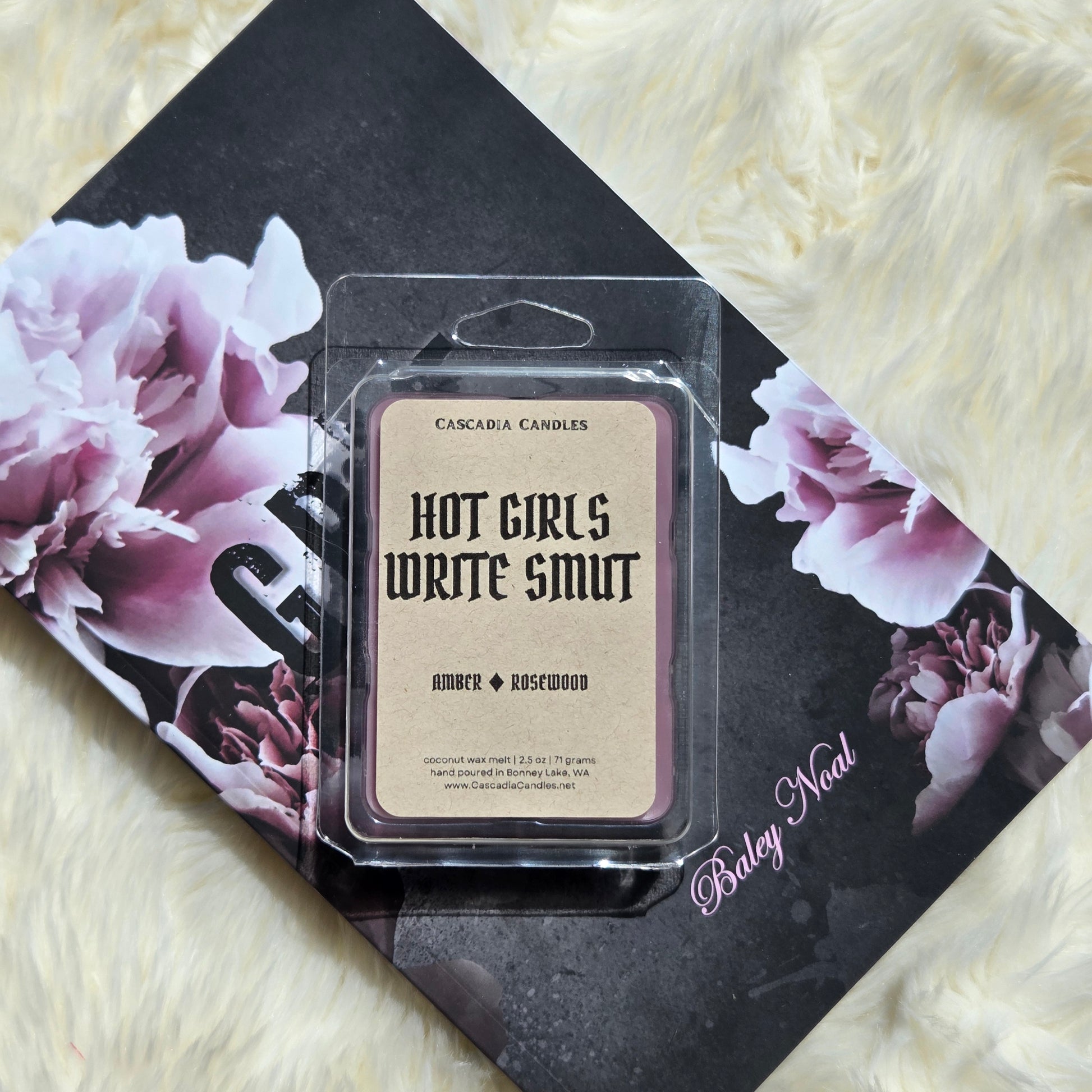 A packaged wax melt product labeled 'HOT GIRLS WRITE SMUT' by Cascadia Candles, placed on top of a black floral book against a cream faux-fur surface. The product is in a clear plastic clamshell with a Kraft label and purple wax visible around the edges.