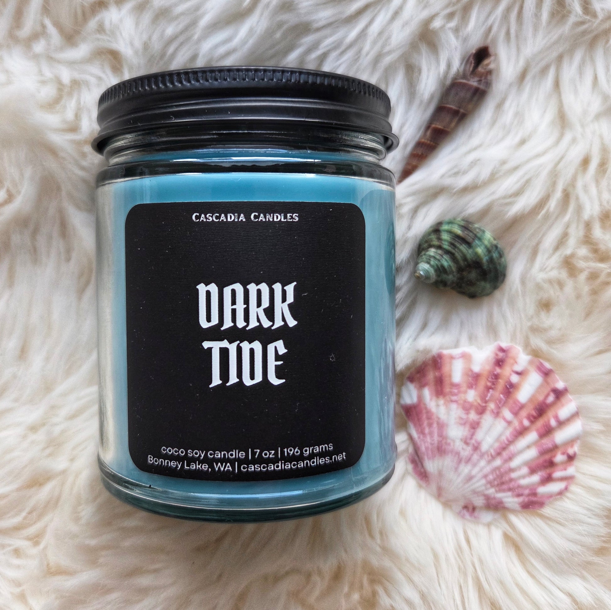 Candle labeled 'Dark Tide' by Cascadia Candles on a fluffy white surface with shells.