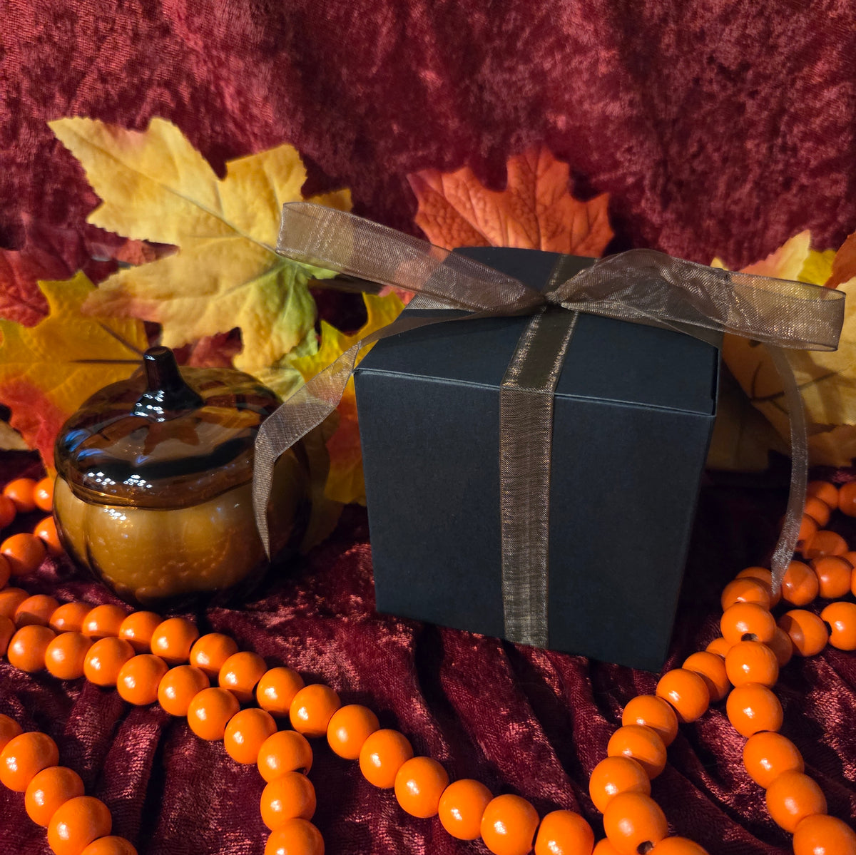 A square black gift box tied with a sheer brown ribbon, styled with orange beads, a small amber glass pumpkin candle, and autumn leaves against a dark red velvet background.