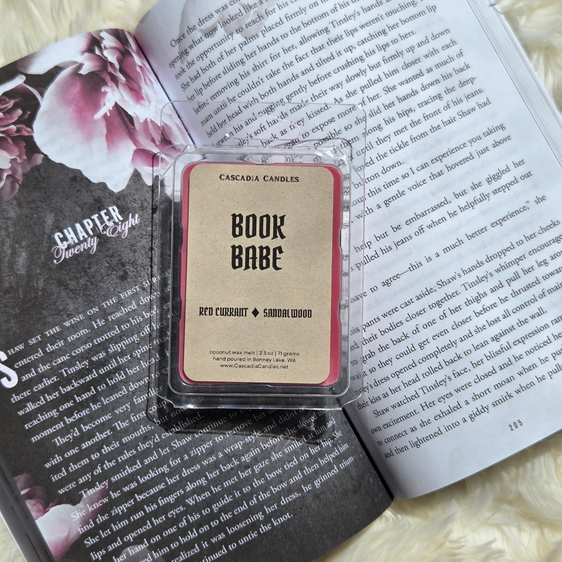 A clear clamshell wax melt package labeled 'Cascadia Candles Book Babe' sits on top of an open book with printed text and a floral page. The wax melt has a Kraft label with black text.