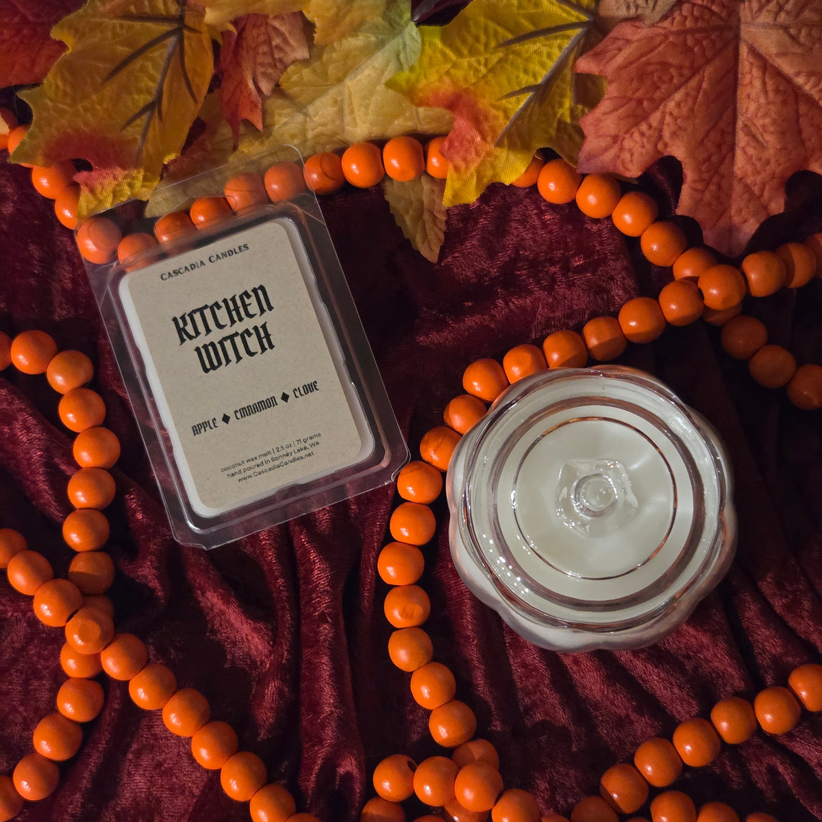 A packaged wax melt labeled 'Kitchen Witch' by Cascadia Candles, shown next to an glass jar pumpkin-shaped candle with white wax, styled on a dark burgundy velvet fabric with orange beads and autumn leaves.