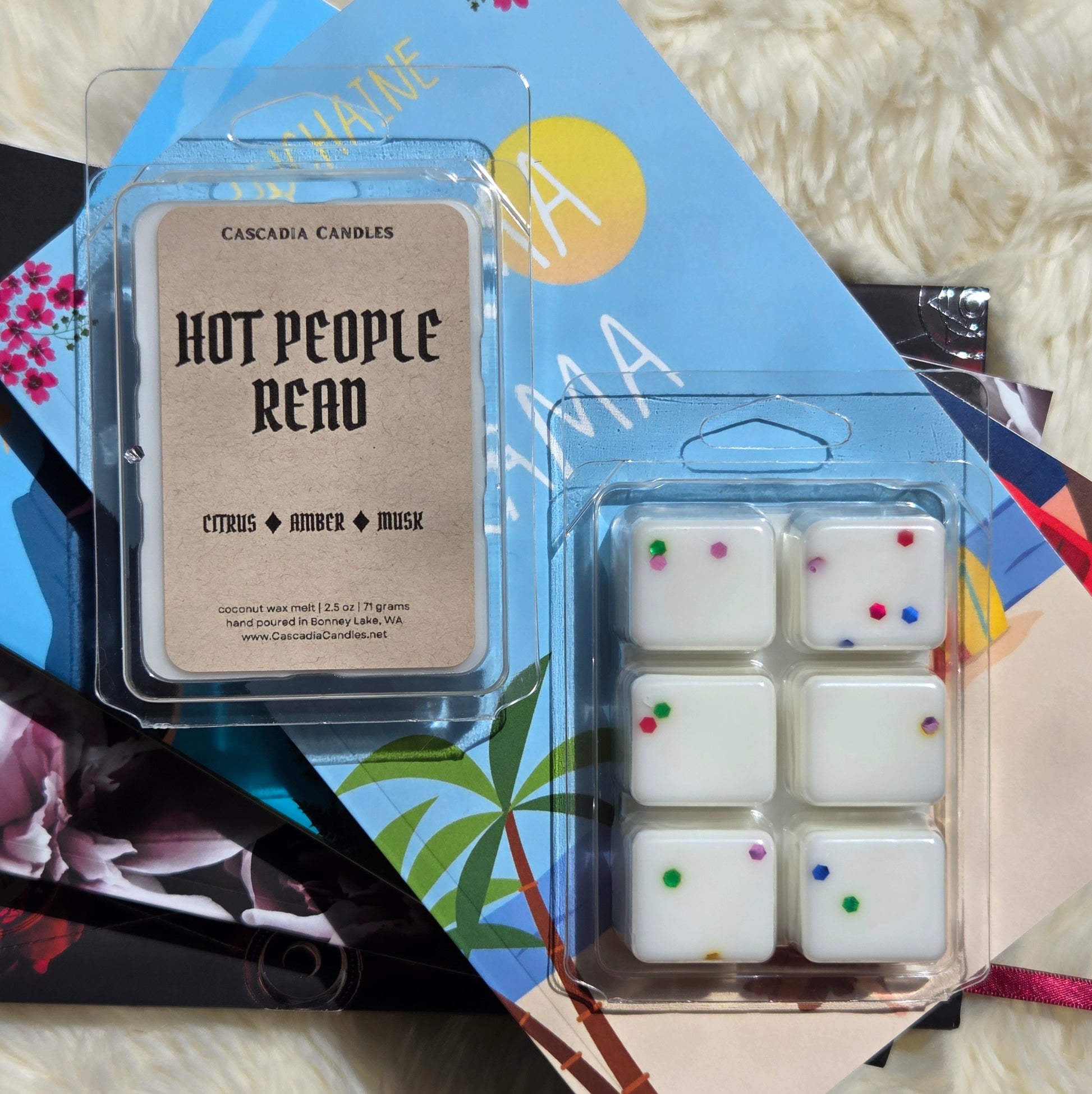Two packaged scented wax melts from Cascadia Candles, including the 'Hot People Read' citrus and amber melt on the left and a six-piece white melt set with colorful eco-friendly glitter on the right, styled on top of a tropical-themed book over a white faux fur background.