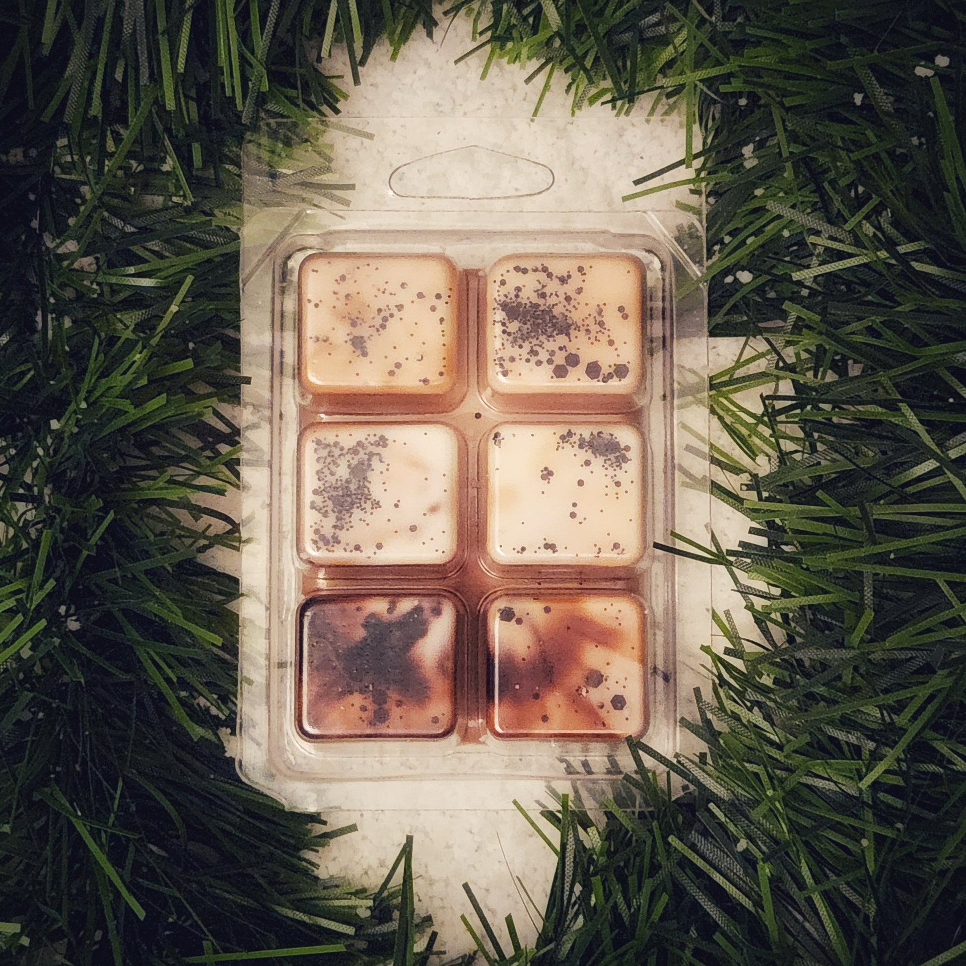 Pack of six square wax melts in a clear plastic packaging on a bed of green garland and snow.