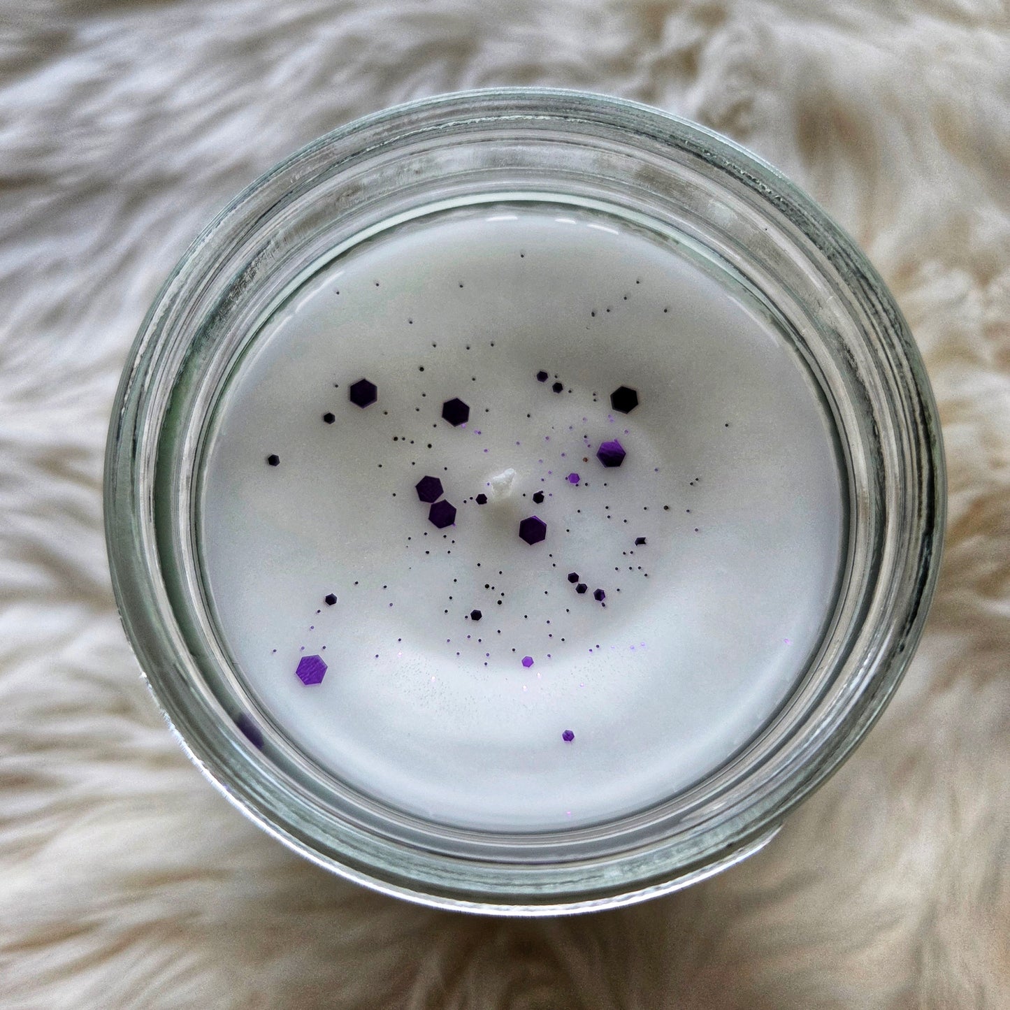 Glass jar with white candle inside, sprinkled with purple glitter on a fluffy white surface.