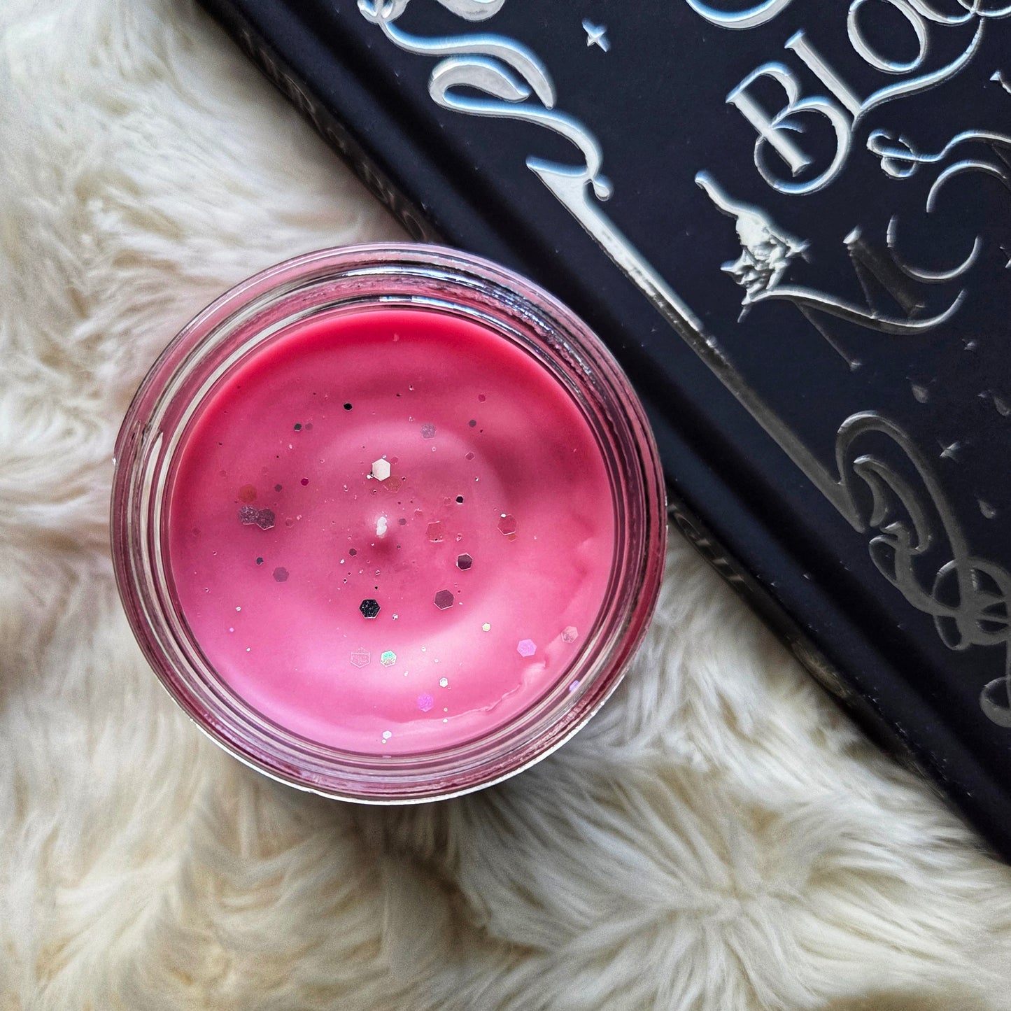Pink glittery cream in a jar on a fluffy white surface with a black and silver patterned book.