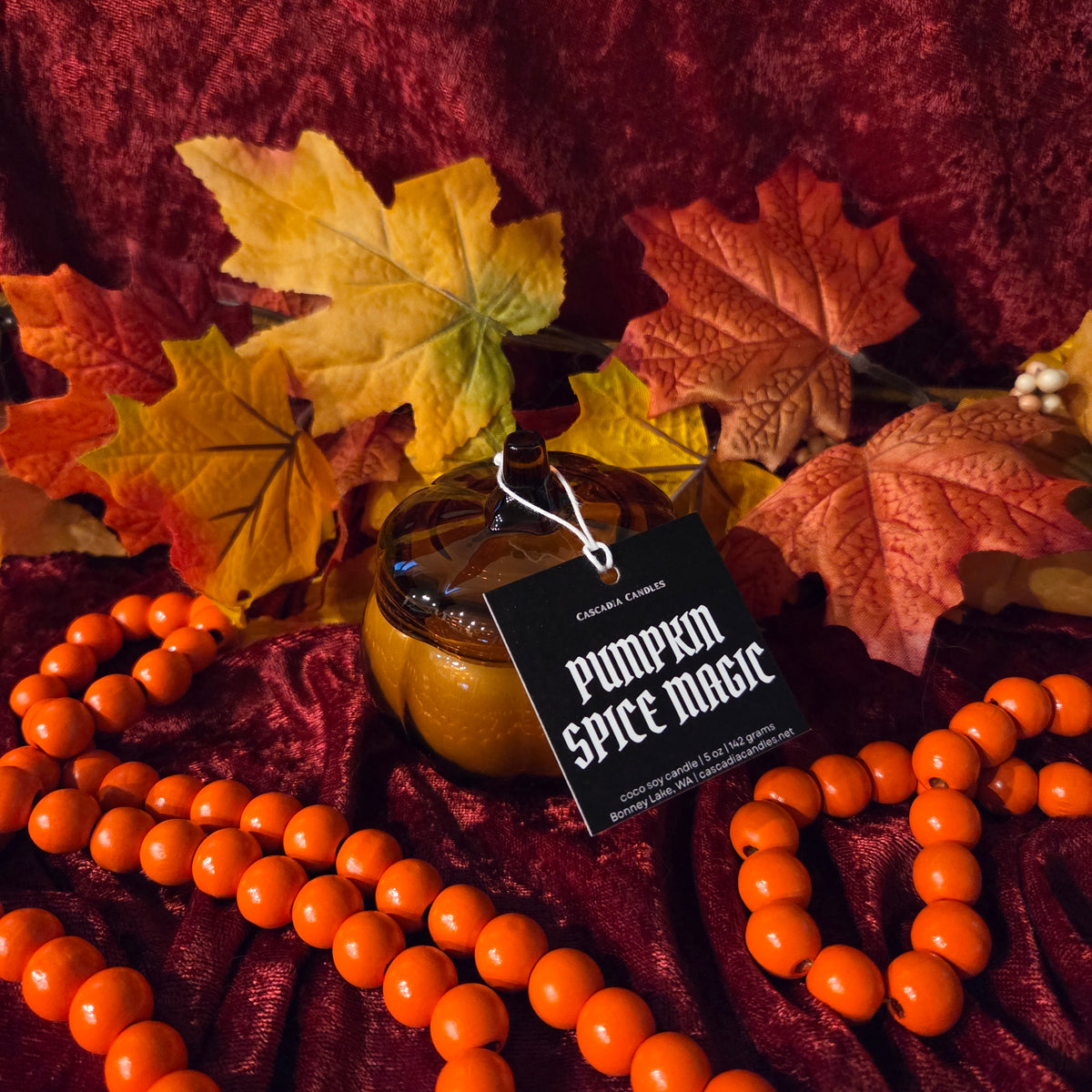 A small glass pumpkin-shaped jar candle with a lid sits on dark red fabric surrounded by autumn leaves and orange beaded garland. A black hanging tag reads 'Pumpkin Spice Magic' and shows the Cascadia Candles brand.