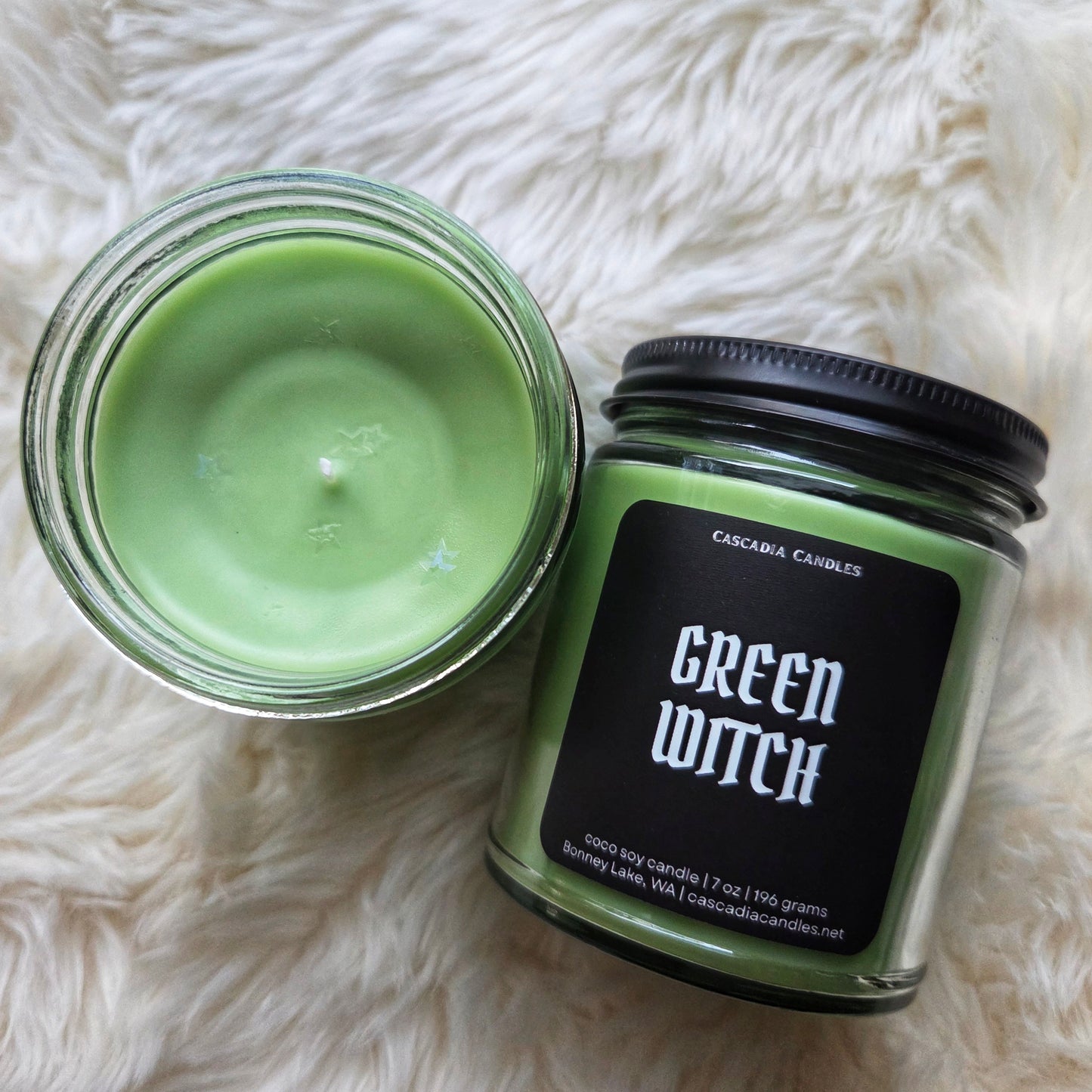 Green candle in a glass jar with a black lid labeled 'Green Witch' on a white fluffy surface.