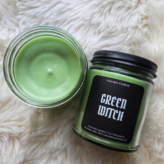 Green candle in a glass jar with a black lid labeled 'Green Witch' on a white fluffy surface.