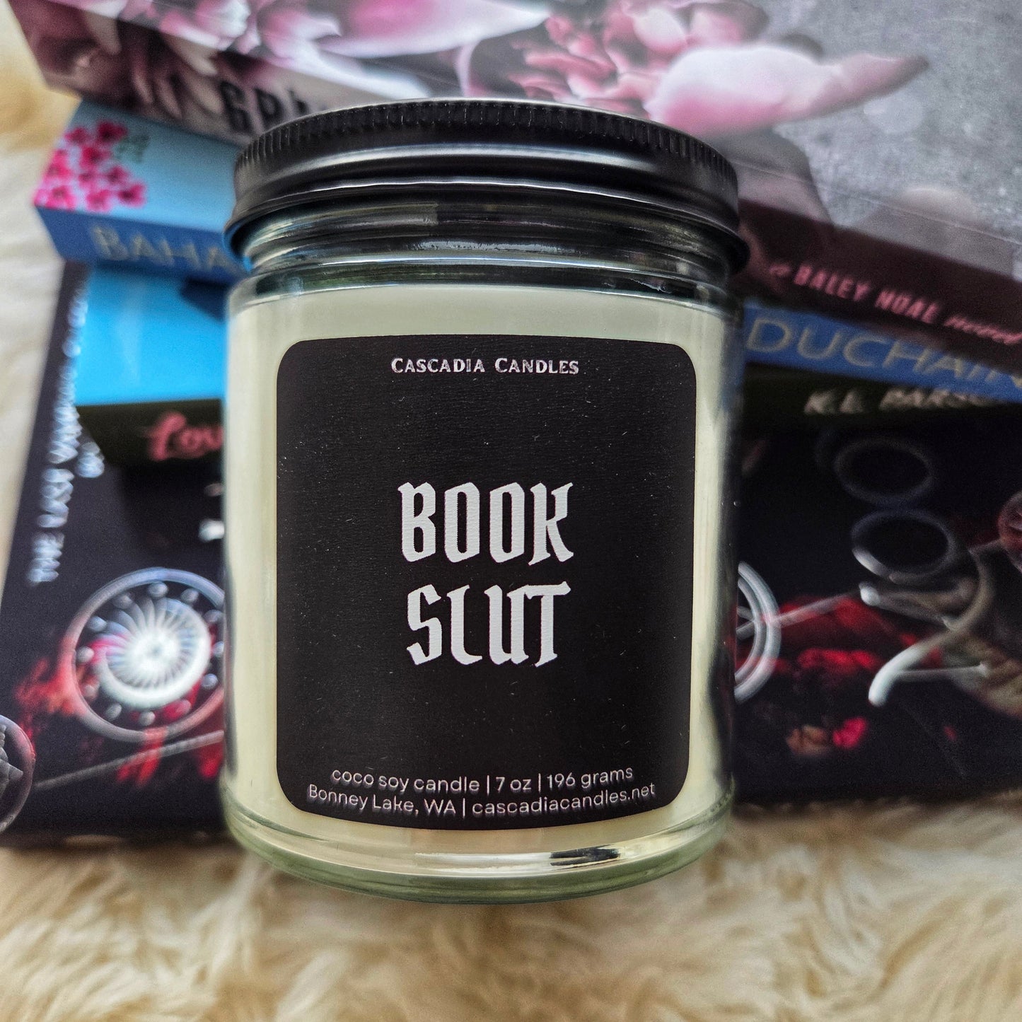 Candle jar labeled 'Book Slut' by Cascadia Candles on a surface with books in the background.