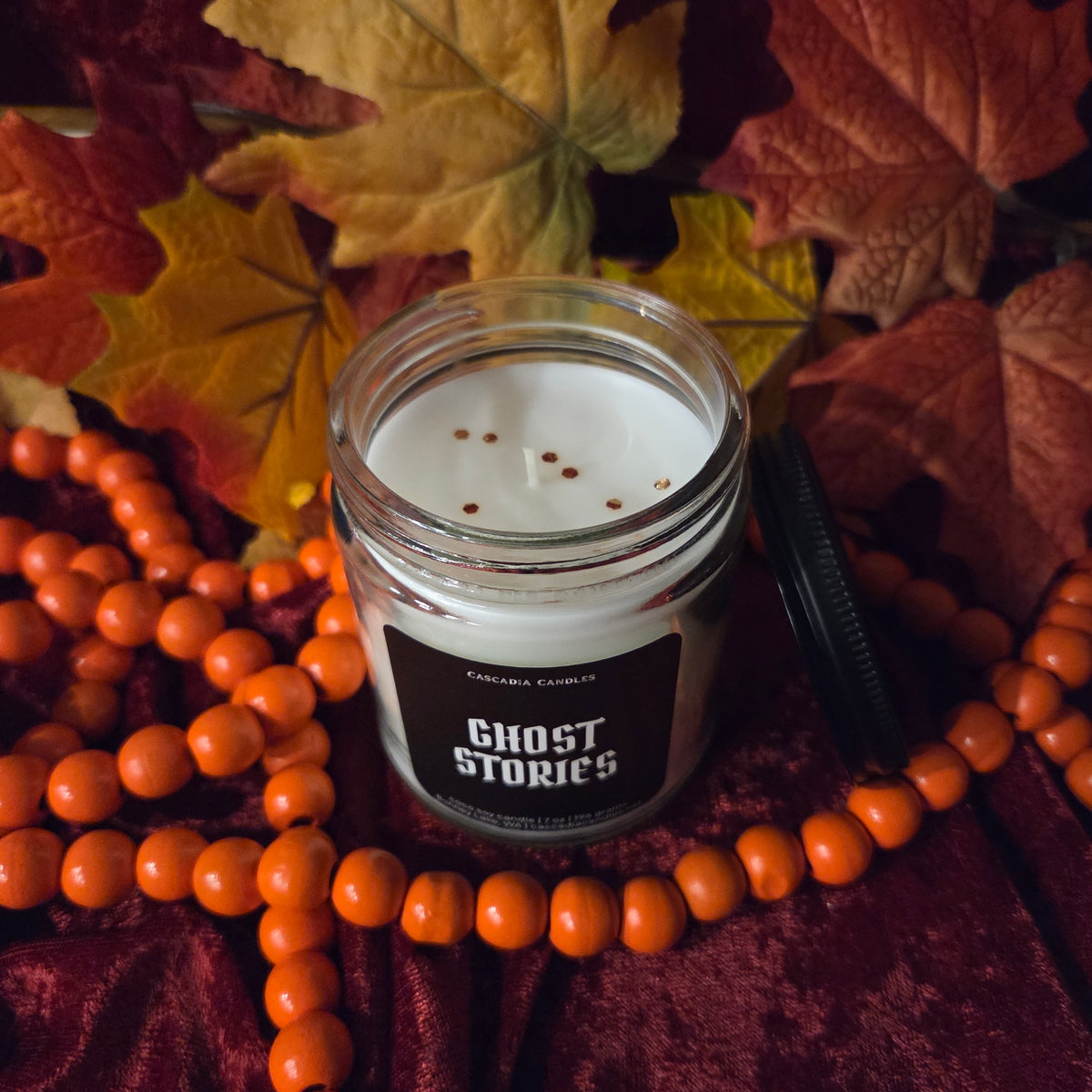 A glass jar candle labeled 'GHOST STORIES' by Cascadia Candles, styled with orange beads and autumn leaves on a dark red fabric background.